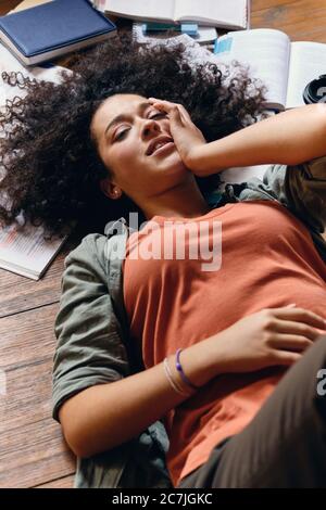 Giovane studentessa con capelli ricci scuri che si trova sul pavimento con libri di testo intorno e occhi di chiusura stancanti a casa Foto Stock