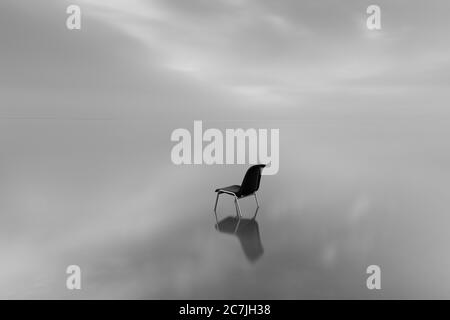 Immagine in scala di grigi di una sedia su una superficie d'acqua con una riflessione su un giorno piovoso Foto Stock