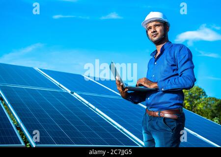 Giovane ingegnere maschile con computer portatile in piedi a mano vicino pannelli solari, terra agricola con cielo azzurro chiaro sfondo, energia rinnovabile, ener pulito Foto Stock