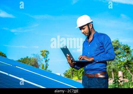 Giovane ingegnere maschile con computer portatile in piedi a mano vicino pannelli solari, terra agricola con cielo azzurro chiaro sfondo, energia rinnovabile, ener pulito Foto Stock