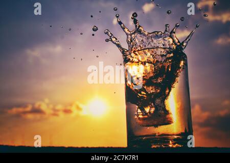 Cubo di ghiaccio in un bicchiere di alcol formando uno spruzzi con il tramonto sullo sfondo Foto Stock