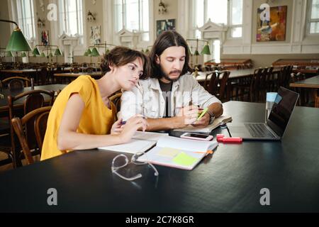 Due giovani studenti pensivi che fanno appunti mentre studiano con il laptop insieme in biblioteca di università Foto Stock