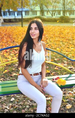 Bella ragazza con i capelli scuri si siede su una panca del parco, sorriso felice, giorno di sole autunno, foglie gialle su alberi e terra, primo piano Foto Stock