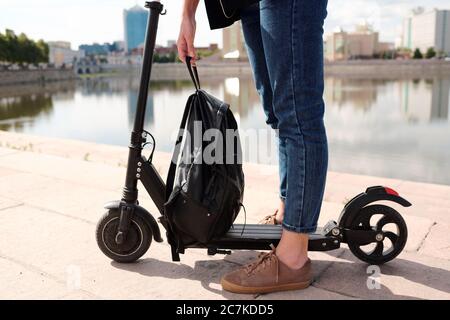 Gambe di giovane donna in jeans blu che tengono zaino in pelle nera sopra scooter elettrico mentre si levano in piedi contro il fiume e il paesaggio urbano Foto Stock