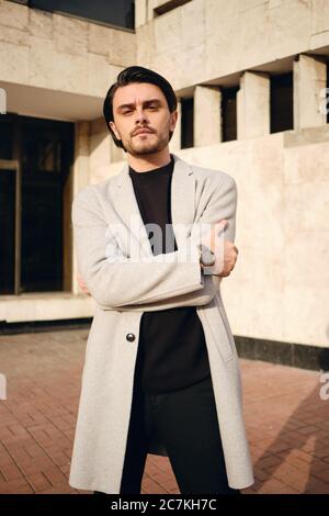 Giovane bell'uomo serio in giacca con mani incrociate che guarda con sicurezza in macchina fotografica all'aperto Foto Stock