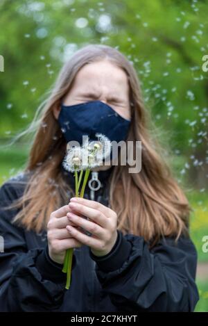 Scolastica con maschera contro corona, tenendo dandelioni, semi che volano attraverso l'aria Foto Stock
