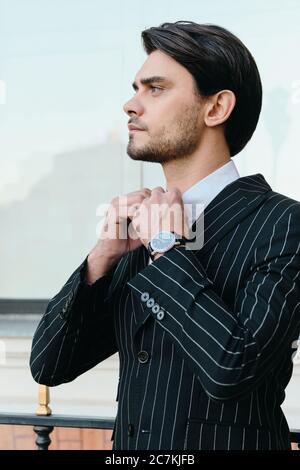 Giovane bel bruna con cuscinetti in camicia bianca e giacca classica a righe che guarda da parte mentre si trova in città da sola Foto Stock