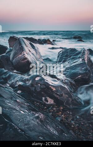 onde che si infrangono su rocce bagnate sulla costa durante il tramonto Foto Stock