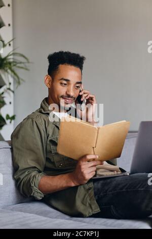 Giovane attraente uomo afroamericano sorridente con notebook che parla felicemente sul cellulare in spazio co-working Foto Stock
