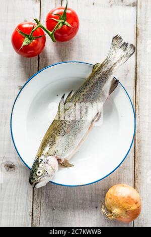 Trota fresca dal mercato su un piatto marittimo con due pomodori rossi e una cipolla su fondo bianco di legno Foto Stock