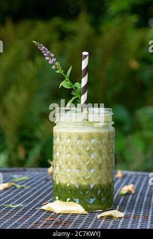 Frullato verde in un bicchiere con fiori di paglia e menta su tavolo da giardino, sfondo sfocato, verde. Foto Stock