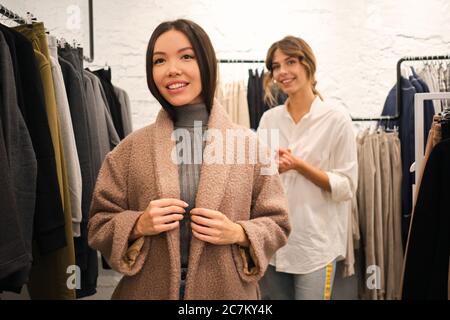 Bella ragazza asiatica di stile gioiosamente cercando su nuovo cappotto casual in negozio di vestiti Foto Stock