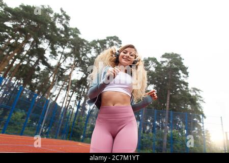 Donna blonde sorridente in rosa leggins, che si gode di jog workout sul campo rosso, correre in cuffie, ascoltare musica. Alberi sfocati e. Foto Stock