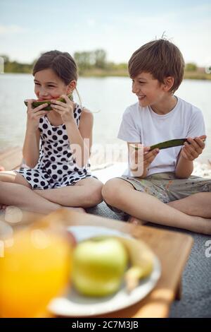 Ragazza e ragazzo carino che mangiano cocomelon fresco sul molo. I bambini si divertono insieme al lago. Foto Stock