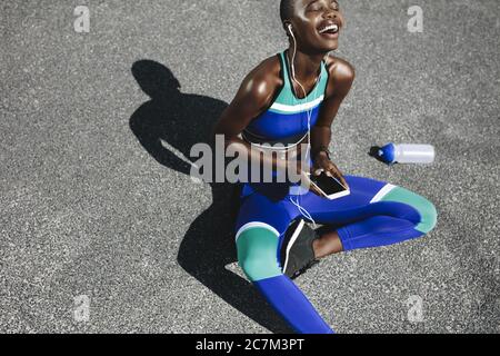 Vista dall'alto della donna africana seduta sulla strada dopo una corsa mattutina e sorridente. Una donna che si prende una pausa dopo un allenamento all'aperto. Foto Stock