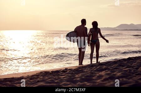 giovane coppia caucasica che cammina sulla spiaggia sabbiosa, portando tavole da surf, parlando Foto Stock