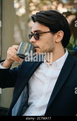 Giovane attraente bruna con bearded uomo in camicia bianca, giacca classica e occhiali da sole che guarda da parte sognando mentre beve caffè sulla strada della città Foto Stock