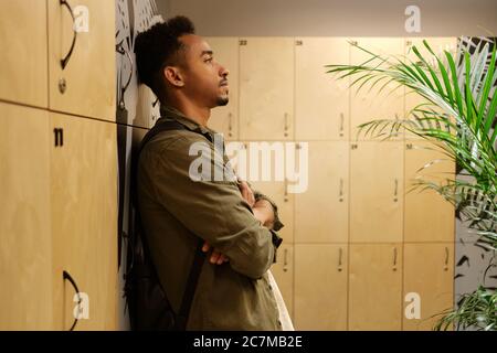 Vista laterale di un giovane e grazioso uomo afro-americano casual, che si trova in piedi pensieroso solo in spogliatoio Foto Stock