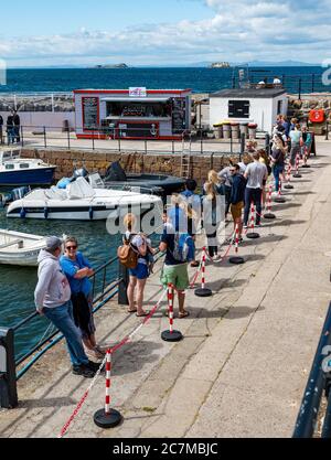 North Berwick, East Lothian, Scozia, Regno Unito, 18 luglio 2020. Regno Unito Meteo: Sole estivo in una città molto trafficata di mare che è tornato alla normalità, anche se con misure di allontanamento sociale in atto. Il popolare Lobster Shack ha una lunga coda socialmente distanziata intorno al porto Foto Stock