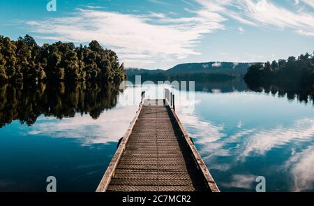 Percorso in legno con alberi e un cielo blu riflesso Lago Mapourika Waiho in Nuova Zelanda Foto Stock