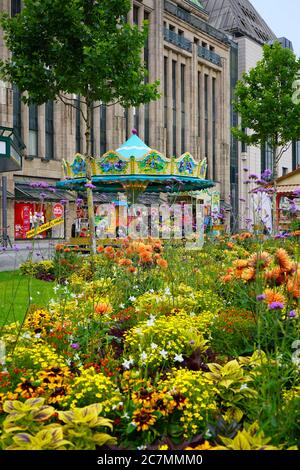 Giostra nostalgica di fronte all'edificio storico del grande magazzino "Kaufhof" con colorati fiori estivi in primo piano. Foto Stock