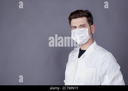 Giovane medico di sesso maschile indossando un abito medico bianco e un maschera facciale bianca Foto Stock