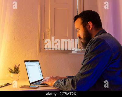 Uomo bearded seduto e digitando qualcosa sul suo laptop Foto Stock