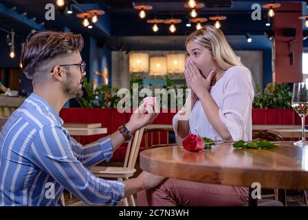 Giovane coppia romantica in un bel ristorante di fantasia. Il ragazzo sorprende la sua ragazza con una rosa rossa e le propone con un anello di fidanzamento. Foto Stock