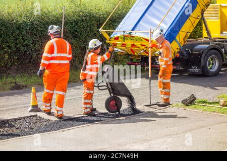 Operai e donne che fanno riparazioni stradali riparando la superficie stradale irregolare danneggiata e buchi di pentola con asfalto in una strada di paese di Cheshire Inghilterra UK Foto Stock