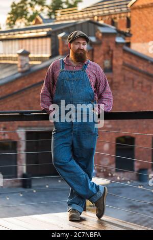 Ritratto di brutale uomo di riparazione motociclistico con cuscinetti blu, camicia e cappello in stile vintage della metà del 20 ° secolo Foto Stock