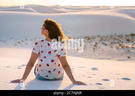 Retro della donna seduta guardando la vista con i capelli nel vento in bianche dune di sabbia monumento nazionale in New Mexico, Stati Uniti al tramonto Foto Stock