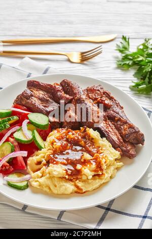 Costolette di maiale servite con purè di patate, salsa di carne, insalata di pomodori tritati, cetrioli, cipolle rosse, prezzemolo su un piatto bianco con c dorata Foto Stock