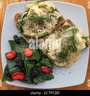 Uova benedettine su fette di pane, salmone, avocado e condite con una salsa hollandaise con alcune erbe aromatiche e accompagnate da uno spinaci Foto Stock