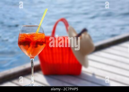 Un bicchiere di cocktail davanti alla borsa intrecciata rossa sfocata con un cappello di paglia e occhiali da sole posti su una passerella in legno vicino al mare con acqua blu. Summe Foto Stock