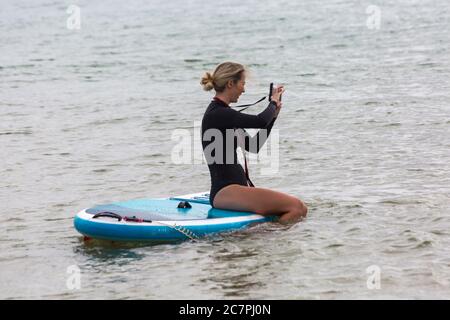 Poole, Dorset UK. 19 luglio 2020. Tempo in Gran Bretagna: Un inizio grigio e frizzante della giornata alle spiagge di Poole. Donna seduta sul paddle board in mare scattando una foto. Credit: Carolyn Jenkins/Alamy Live News Foto Stock