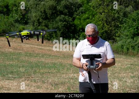 Uomo con capelli grigi che vola un drone su un campo con una tavoletta e una maschera di sicurezza. Nuovo concetto di normalità. Foto Stock