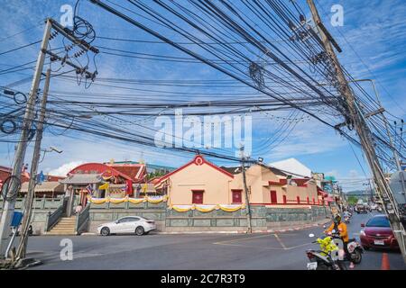 Messily strung i cavi di alimentazione pendono tra i poli di alimentazione su una strada nella città di Phuket, Phuket, Thailandia, con un tempio cinese-taoista sullo sfondo Foto Stock