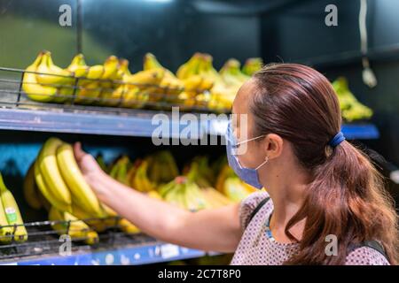 Donna focalizzata in maschera di faccia che sceglie frutta, prendendo banane dagli scaffali nel negozio di alimentari. Cliente in supermercato. Vista laterale. Shopping durante l'epidemia Foto Stock