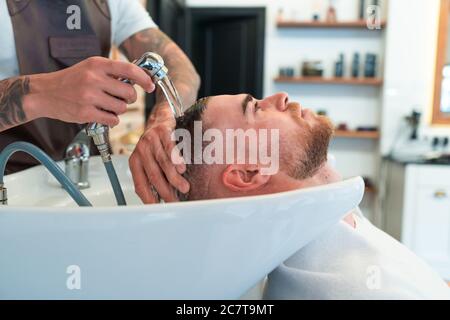Primo piano di un parrucchiere con tatuaggi che lavano i capelli di un giovane cliente maschile Foto Stock