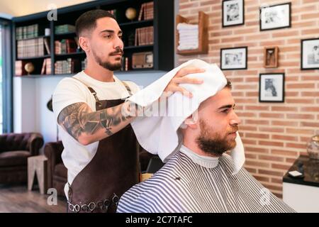 Giovane parrucchiere che asciuga i capelli del cliente maschile con un bianco asciugamano in un barbiere Foto Stock