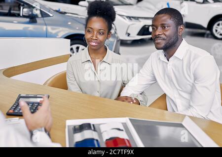 giovane agente di vendita professionale caucasico e clienti afro-americani hanno una conversazione amichevole in concessionaria, i clienti sono venuti a comprare una nuova auto Foto Stock