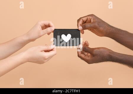 Mani di donna caucasica e di uomo afroamericano che tengono foglio di carta con cuore disegnato su sfondo a colori. Concetto di razzismo Foto Stock