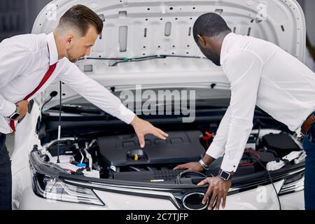 cliente africano e venditore caucasico hanno aperto il cofano dell'auto e discusso tutte le sfumature riguardanti l'auto. in concessionaria Foto Stock