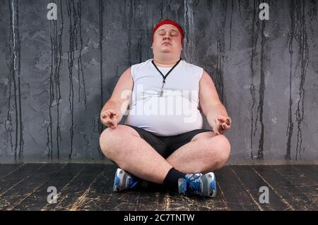 giovane uomo caucasico grasso medita sul pavimento, facendo yoga, si siede in posa yoga. isolato sfondo grigio Foto Stock