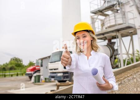 Donna caucasica che indossa un elmetto giallo sorridente e tenente il pollice in cantiere Foto Stock