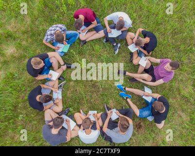 Concetto di lavoro di squadra. Un gruppo di studenti delle scuole superiori si siede sull'erba in un cerchio e si spara in direzioni diverse. Vista drone. Foto Stock