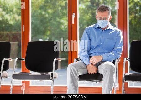 Uomo maturo triste o stanco con maschera di protezione seduta in una sala d'attesa di un ospedale o di un ufficio in una giornata di sole Foto Stock