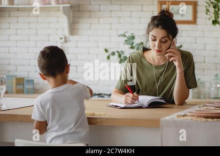 Giovane donna che prende appunti e parla al telefono mentre il suo bambino gioca vicino. Concetto di madre lavoratrice. Foto Stock