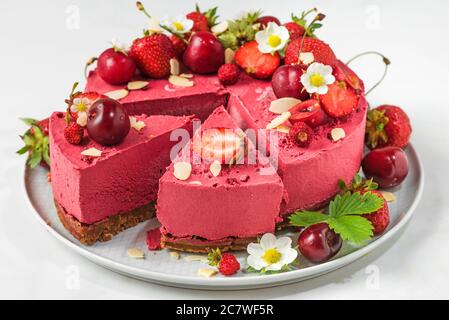 Cheesecake di frutti di bosco con fragole fresche, ciliegie e fiori tagliati in un piatto su sfondo bianco. Primo piano. Dessert con cibo vegano sano Foto Stock