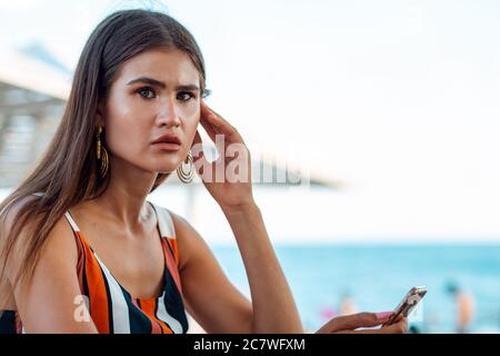 Una donna con un'espressione tesa sta tenendo un telefono nelle sue mani. Caffetteria estiva sulla spiaggia. Concetto di reti sociali e di imprese. Foto Stock